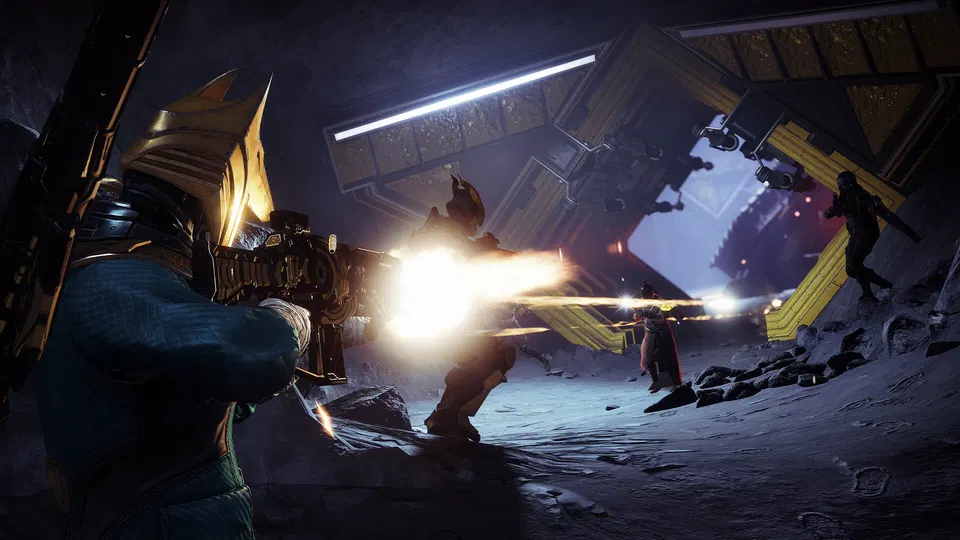 Destiny 2: hráč v zlatě zdobené helmě střílí z těžké zbraně na nepřátele v tmavé hale