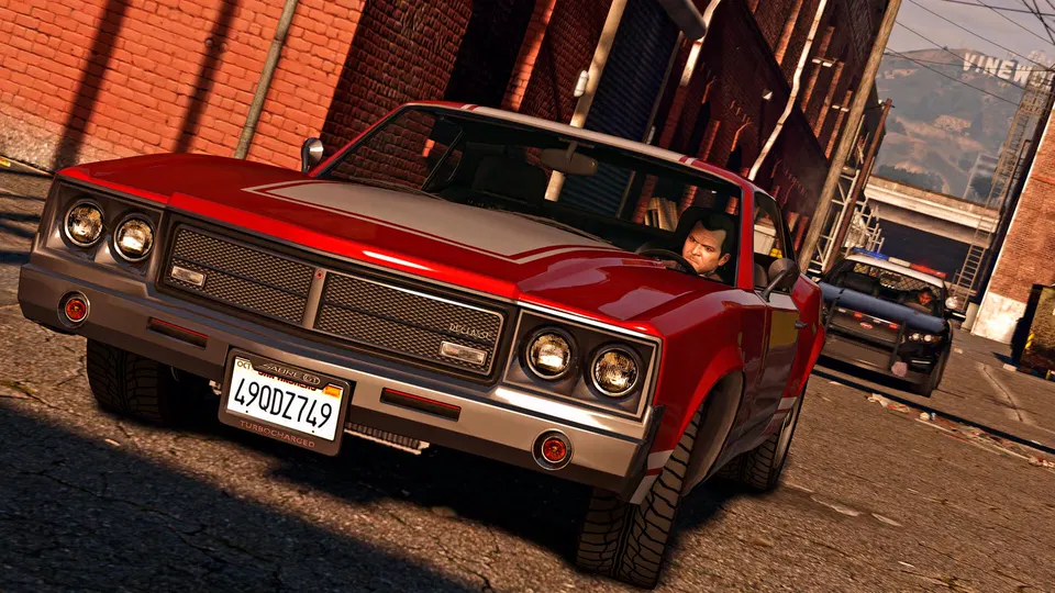 GTA 5: Červený klasický vůz s řidičem jede po městské ulici kolem cihlových budov.