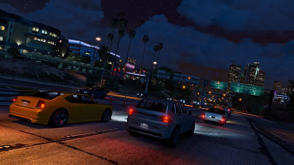 GTA 5: noční silnice v Los Santos s auty na dálnici, palmami a osvícenými městskými budovami v pozadí.