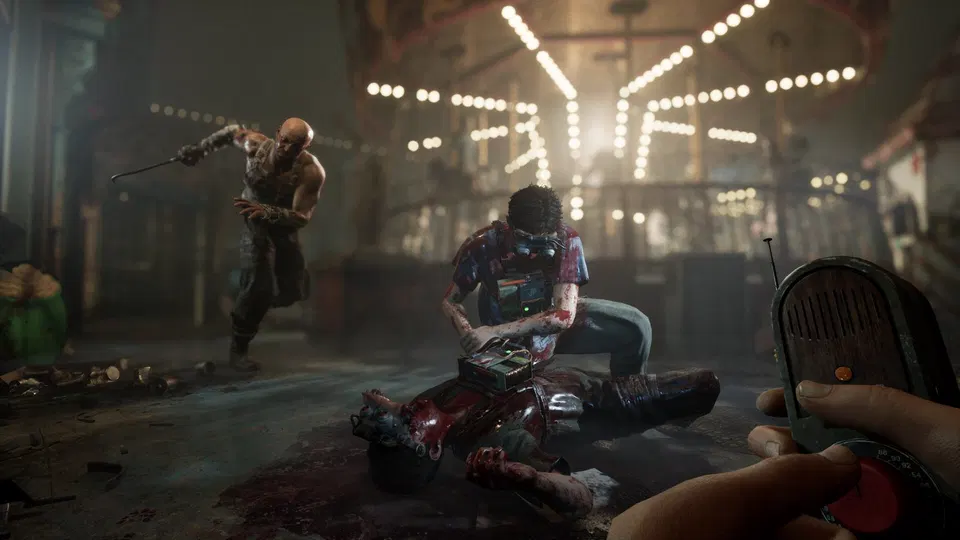 The Outlast Trials: postava klečí u těla v zakrvavené zábavní oblasti s osvětlením, v pozadí další postava s tyčí