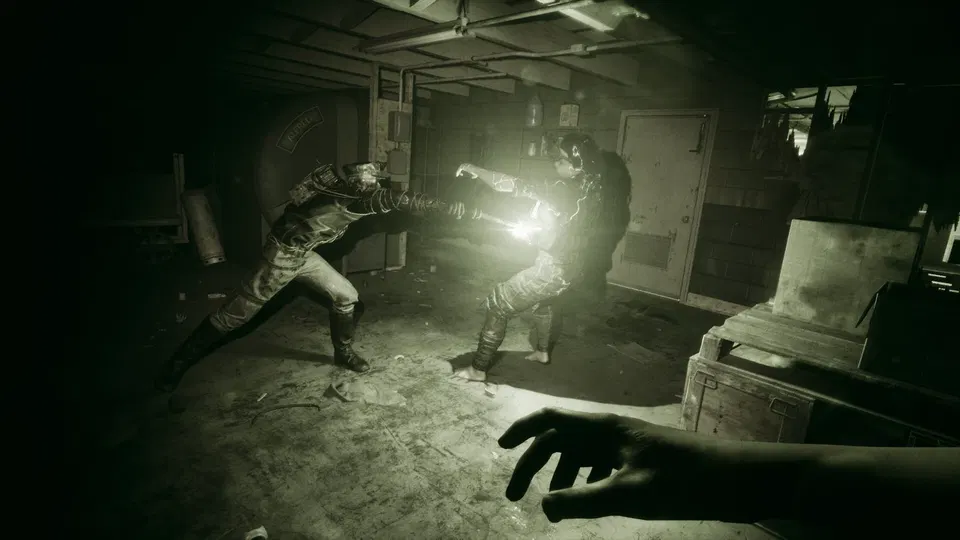 The Outlast Trials: dva lidé se rvou v tmavé místnosti, jeden drží světlo, v popředí ruka hráče